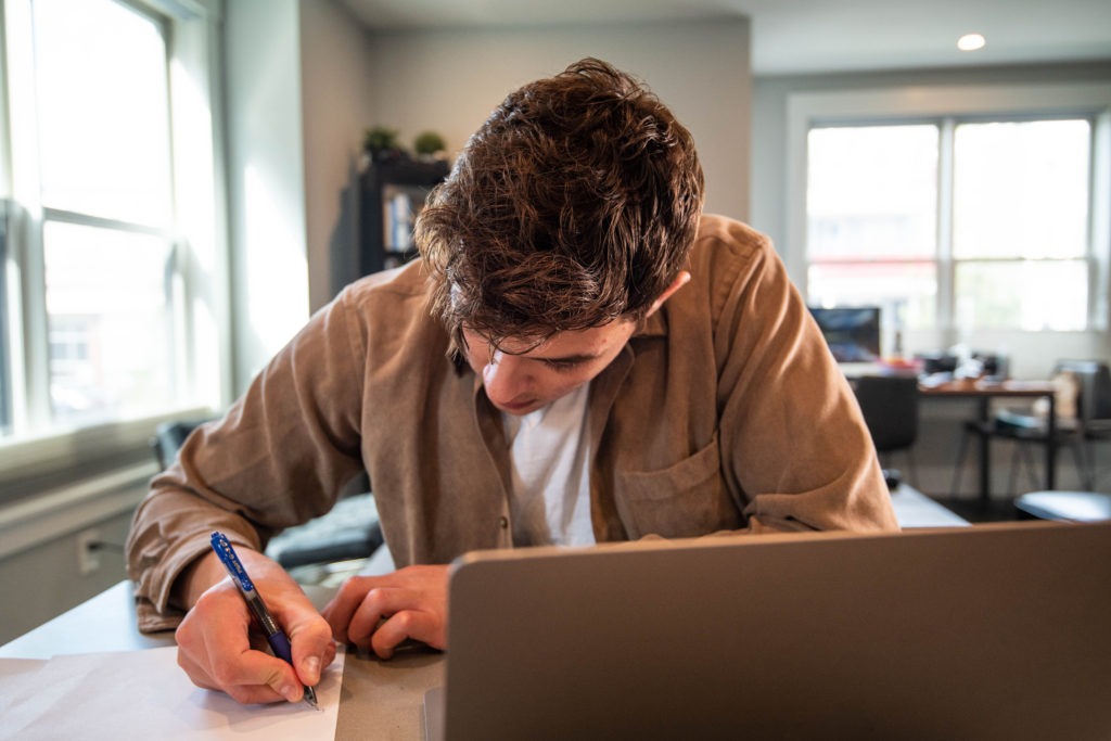 Picture of a young man sitting in front of a laptop while taking notes on paper.